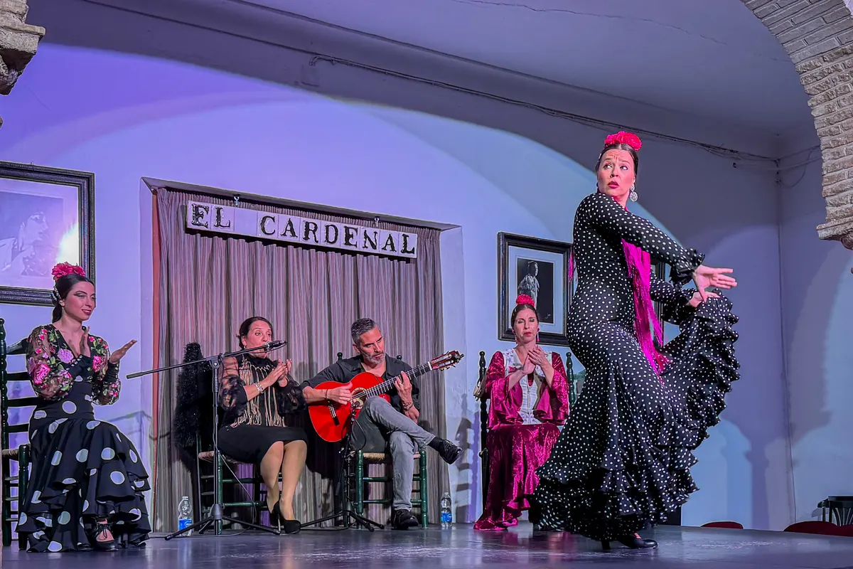 A flamenco dancer in a black polka-dot dress mid-turn at Tablao El Cardenal, with a guitarist, a singer and two seated dancers behind her
