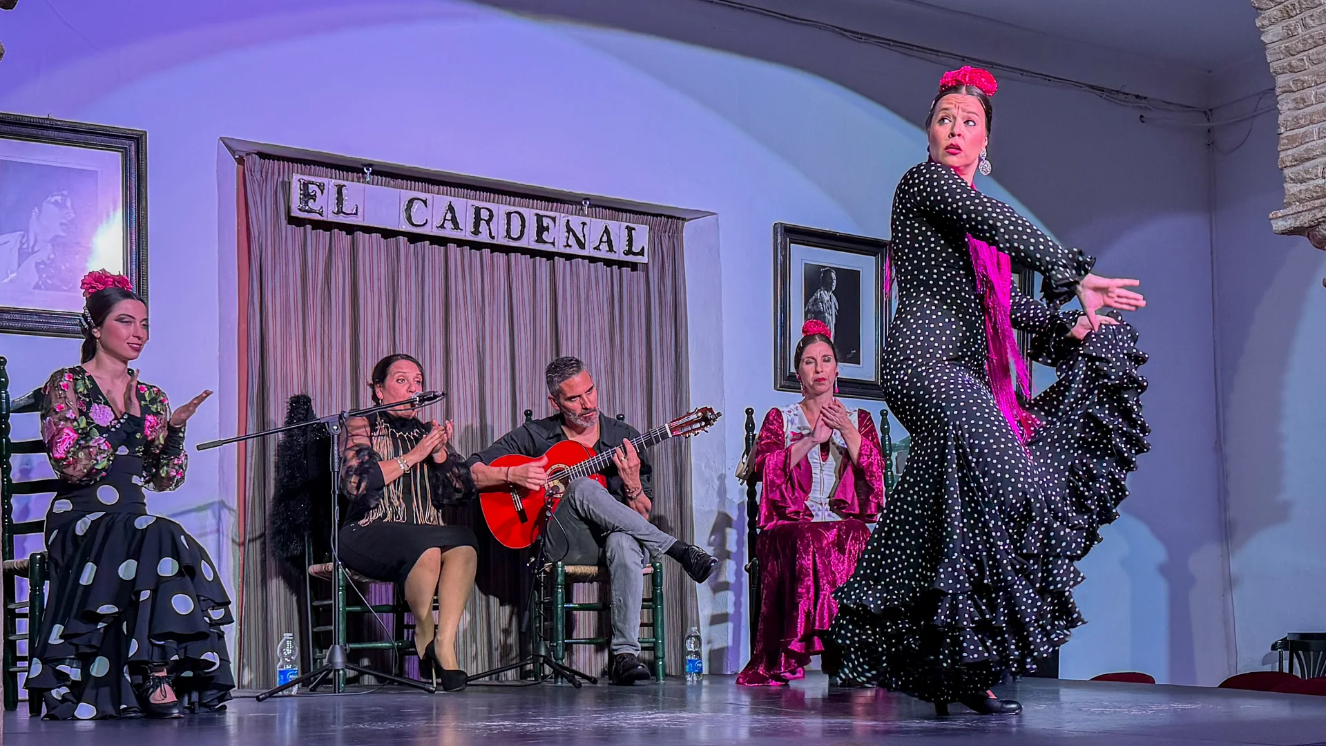 A flamenco dancer in a black polka-dot dress mid-turn on the stage at Tablao El Cardenal, with a guitarist, a singer and two seated dancers behind her under the tiled name sign